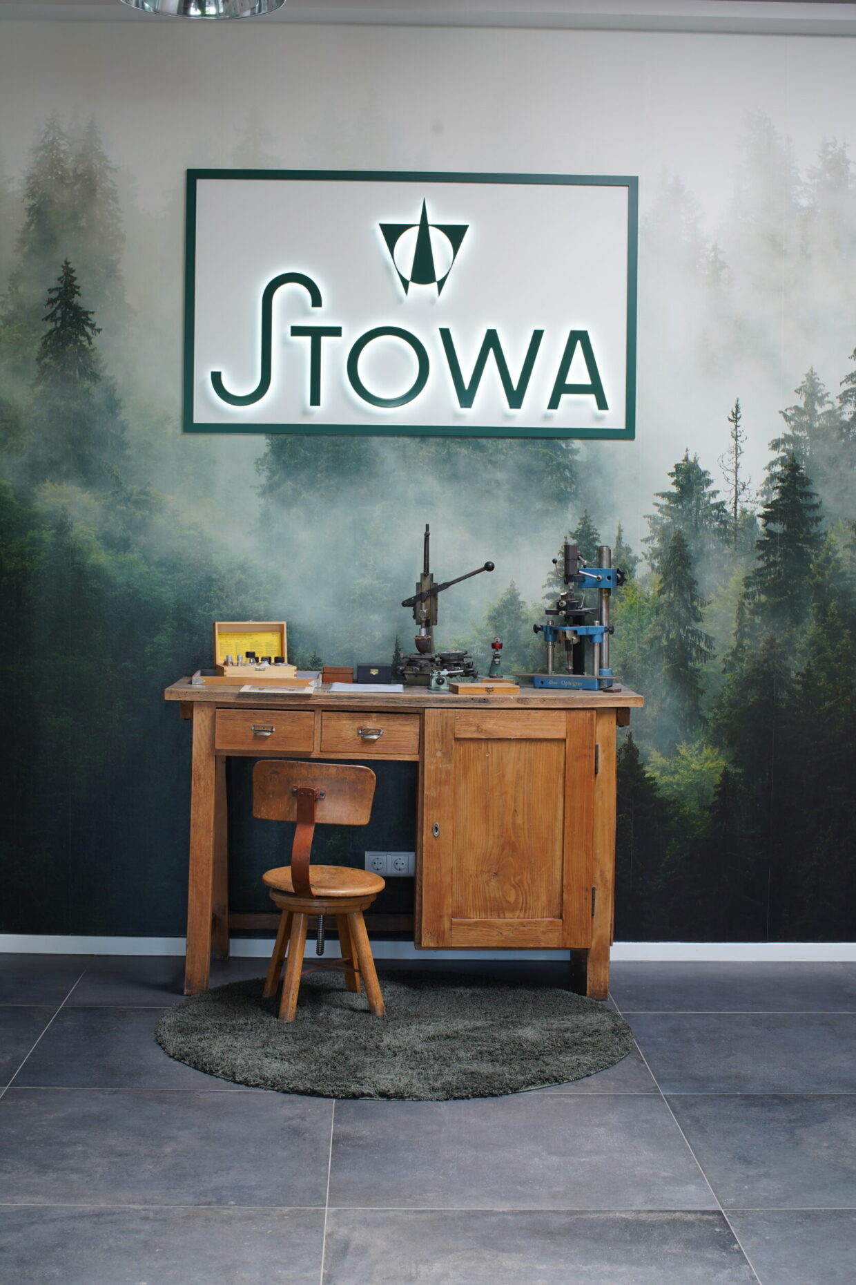 DSC04582-scaled - Deutsche Manufakturenstrasse Holztisch mit Stuhl vor einer Waldtapete, STOWA-Logo über dem Tisch, Werkzeuge und eine kleine Fotografie auf dem Tisch.