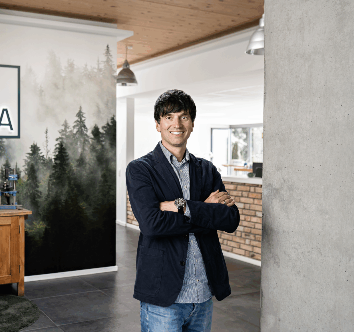 Portrait von Kevin Müller in einem modernen Raum mit Holzdecke und einer Wand mit Waldmotiv. Er steht lächelnd mit versc...