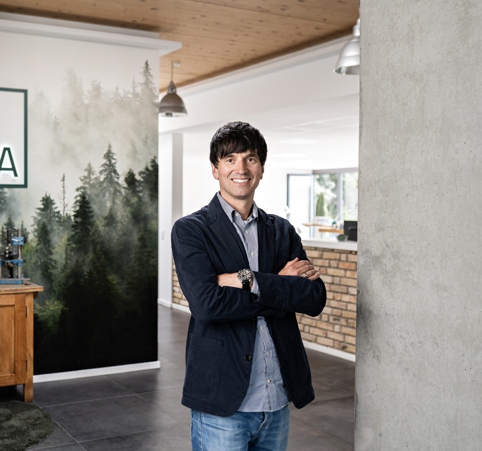 Portrait_Kevin_Mueller_quer_neu - Deutsche Manufakturenstrasse Mann mit dunklen Haaren in einem modernen Raum, lächelt mit verschränkten Armen vor einer Waldtapete.