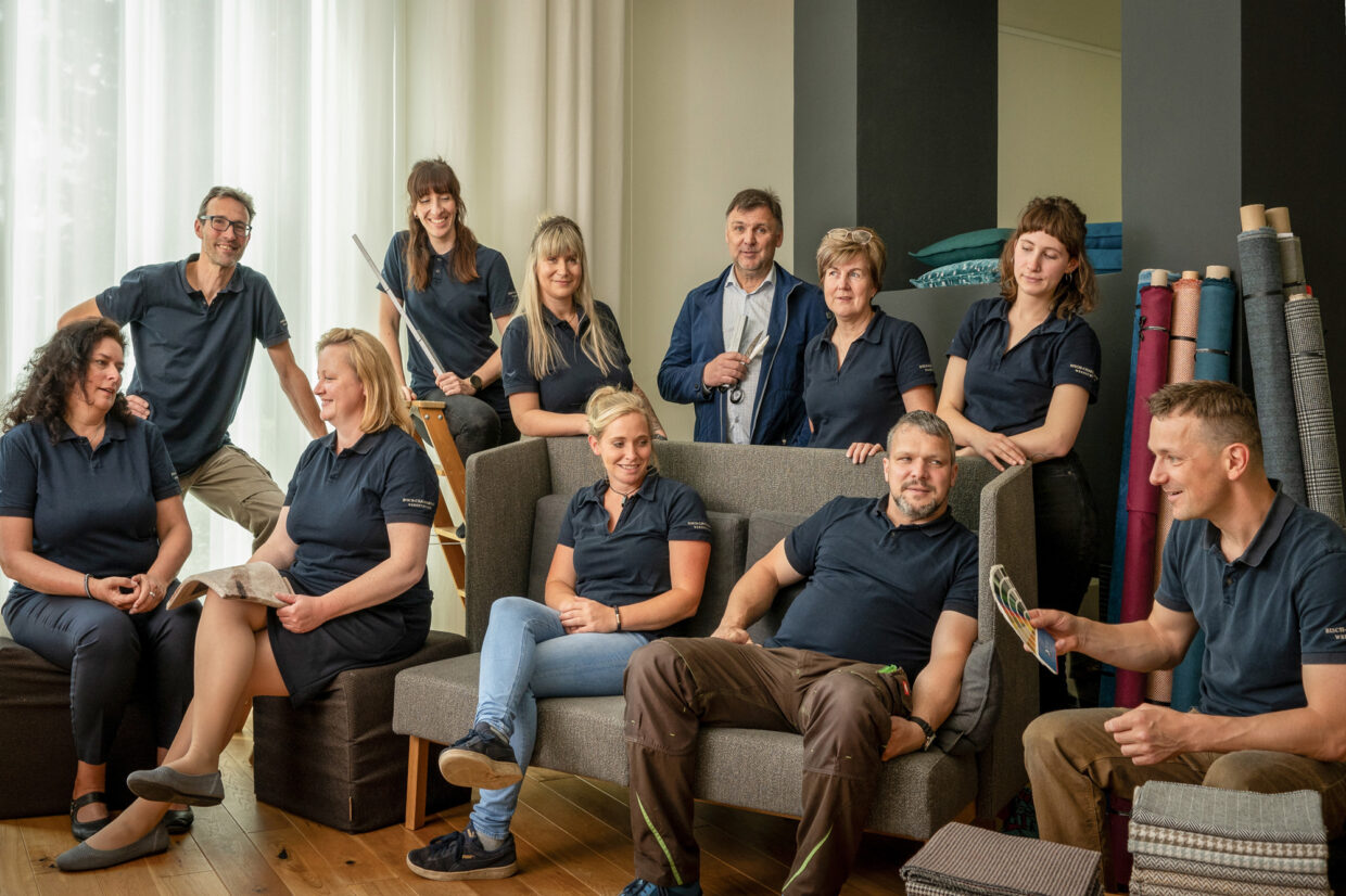 20240606-bc-leger-bearb - Deutsche Manufakturenstrasse Gruppe von zehn Personen in blauen Poloshirts, sitzend und stehend, um ein Sofa in einem hellen Raum, umgeben von Stoffmus...