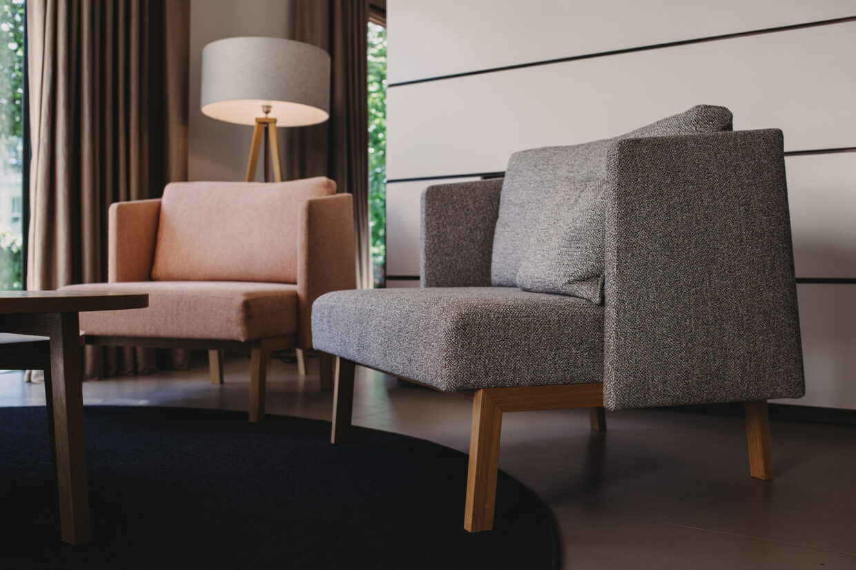 D85_8709 - Deutsche Manufakturenstrasse Zwei moderne Sessel in einem hellen Raum, einer in Grau und einer in Rosa, neben einem runden Tisch und einer Stehlampe.