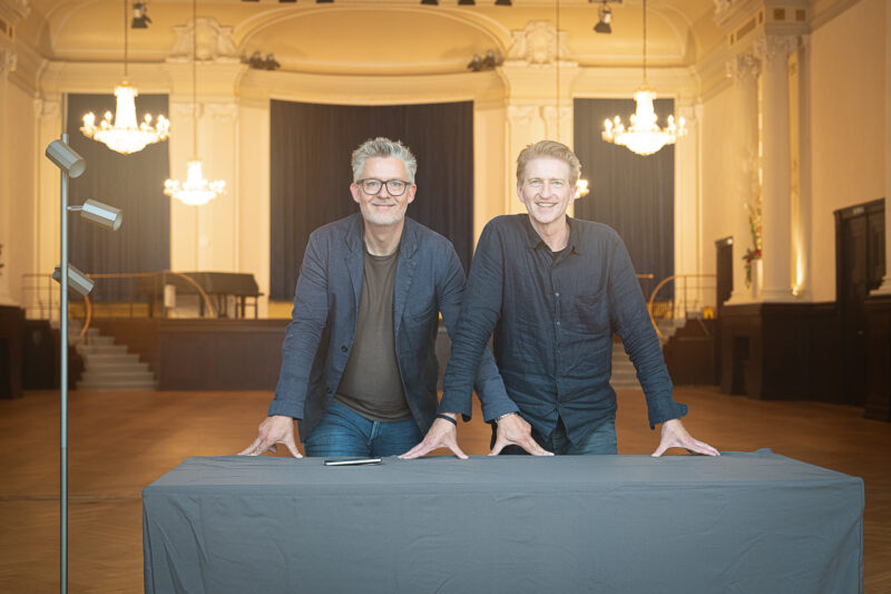 Zwei Männer stehen an einem Tisch in einem eleganten Saal mit Kronleuchtern, im Hintergrund eine Bühne.