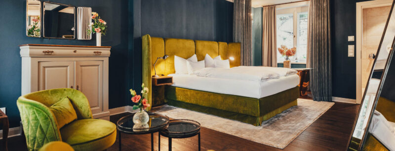 Elegantes Hotelzimmer mit grünem Samtbett, beigem Teppich, stilvollem Sessel und dekorativen Blumenarrangements.