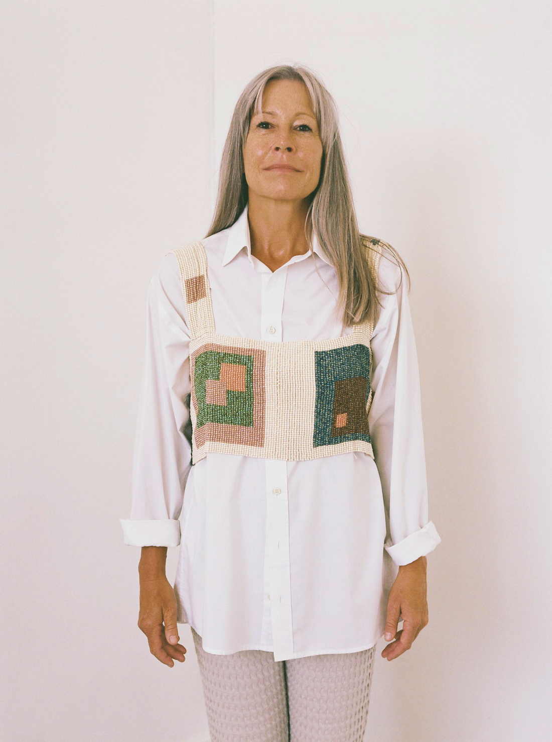 alinanaomi-mom - Deutsche Manufakturenstrasse Frau mit langen grauen Haaren trägt ein weißes Hemd und eine farbige, gestrickte Weste vor neutralem Hintergrund.