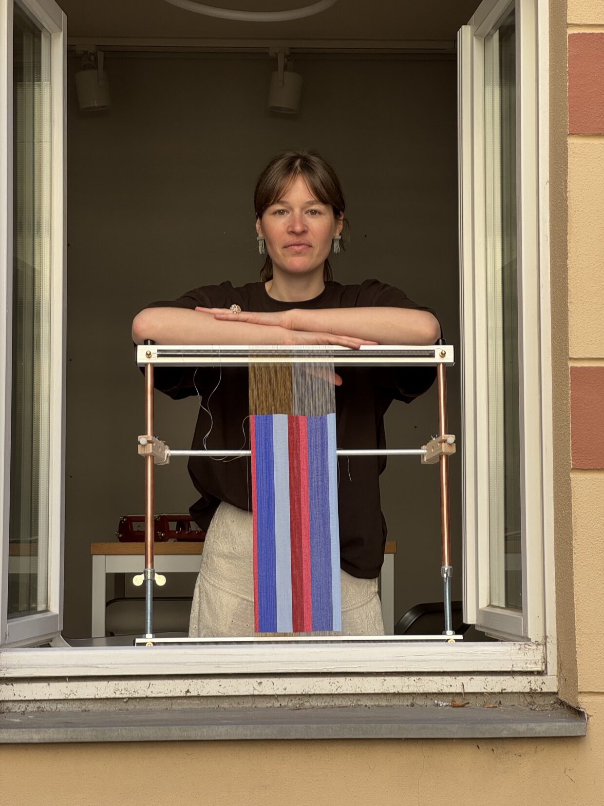 alinanaomi_25 - Deutsche Manufakturenstrasse Frau mit kurzen Haaren steht am Fenster und hält ein Webrahmen mit bunten Fäden.