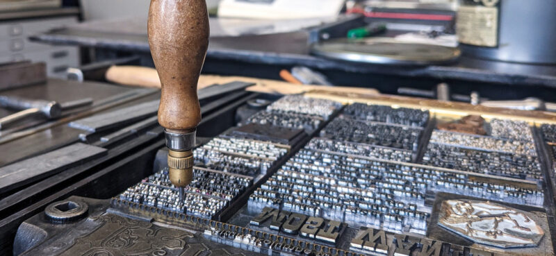 Holzgriff eines Druckwerkzeugs über einer Druckplatte mit feinen Buchstaben und Motiven in einer Druckwerkstatt.