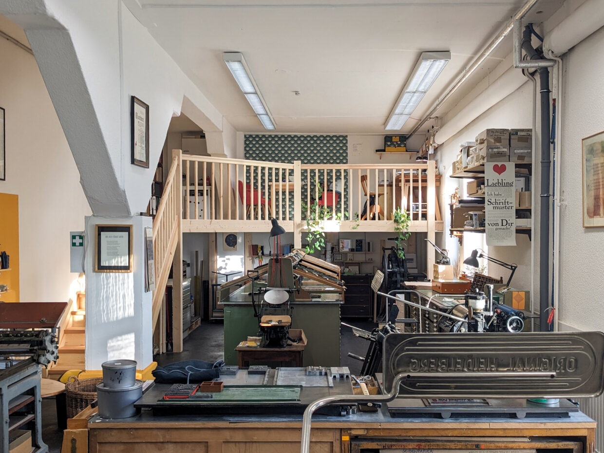 Officina-Arcana---Die-Werkstatt - Deutsche Manufakturenstrasse Helle Werkstatt mit Druckmaschinen, Regalen und einem oberen Bereich, der mit Pflanzen dekoriert ist.