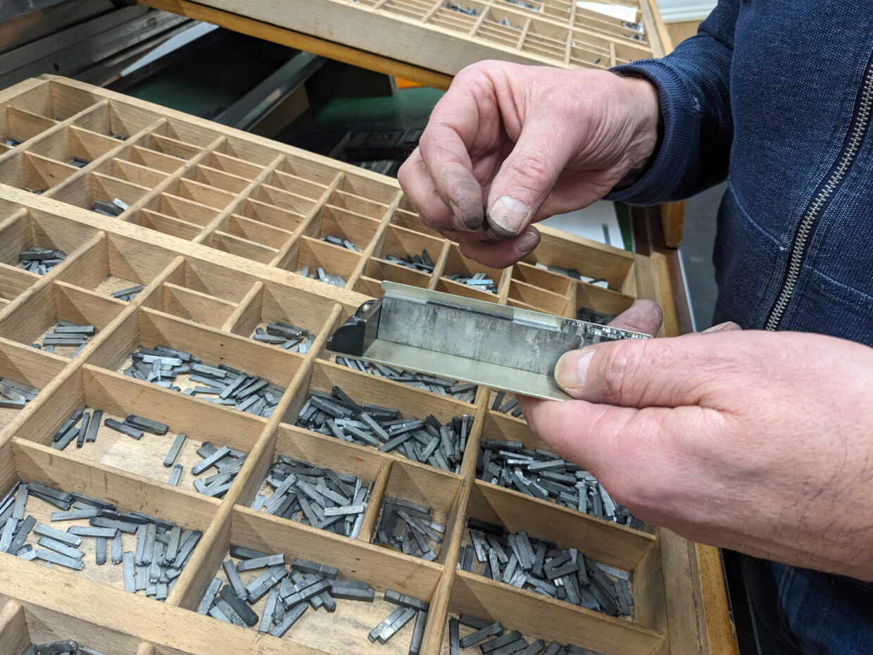 Setzen - Deutsche Manufakturenstrasse Hände halten eine Metallform über einer Holzschublade voller Drucklettern in verschiedenen Größen.