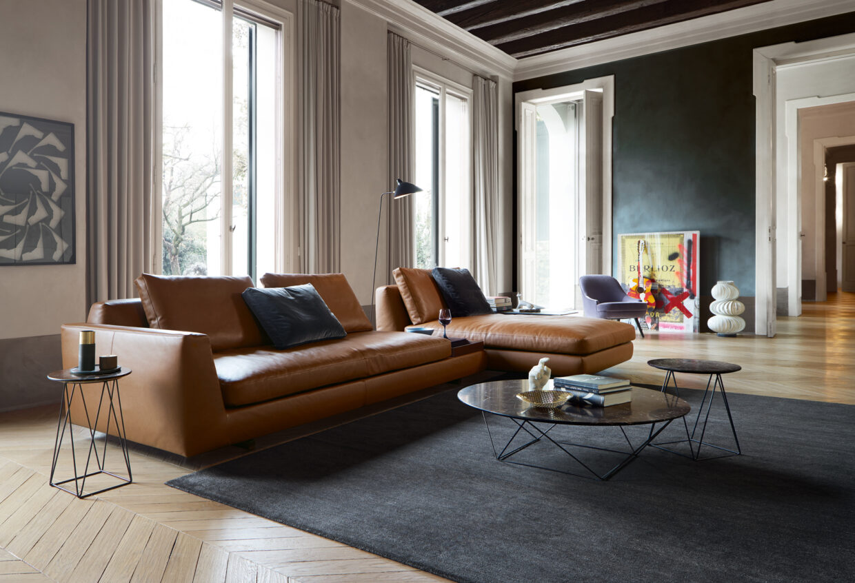 WEBWK-Joco_Stone-0015-H-01_master - Deutsche Manufakturenstrasse Elegantes Wohnzimmer mit braunem Ledersofa, Couchtisch und Kunstwerken an der Wand. Helle Fenster sorgen für Tageslicht.