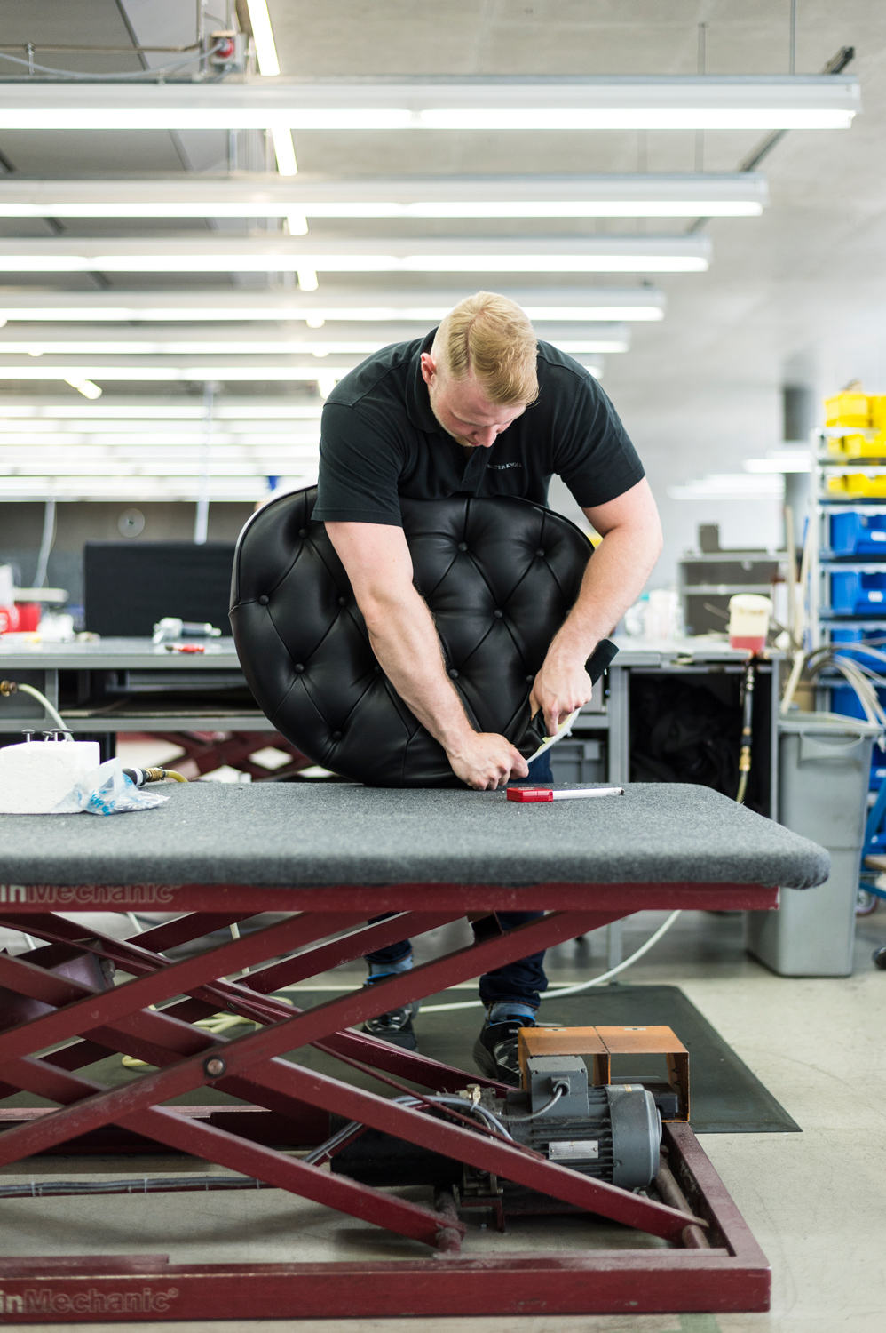 WEBWK_Polsterei-0006_master - Deutsche Manufakturenstrasse Handwerker polstert einen schwarzen, gepolsterten Stuhl auf einem Arbeitstisch in einer modernen Werkstatt.