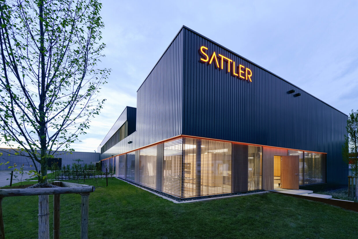 01_SATTLER_Unternehmenssitz_Foto Andreas Körner - Deutsche Manufakturenstrasse Moderne Unternehmenszentrale von Sattler, mit beleuchtetem Schriftzug und großzügigen Fensterfronten, umgeben von Grünf...