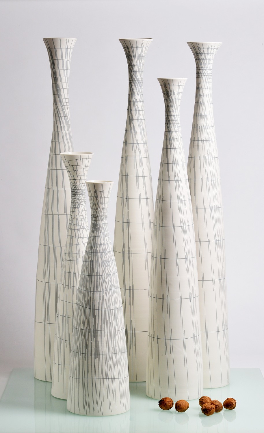 1m Flaschengruppe 1 - Deutsche Manufakturenstrasse Sechs schlanke, weiße Vasen mit feinen grauen Linien, auf einem gläsernen Tisch, umgeben von drei Nüssen.