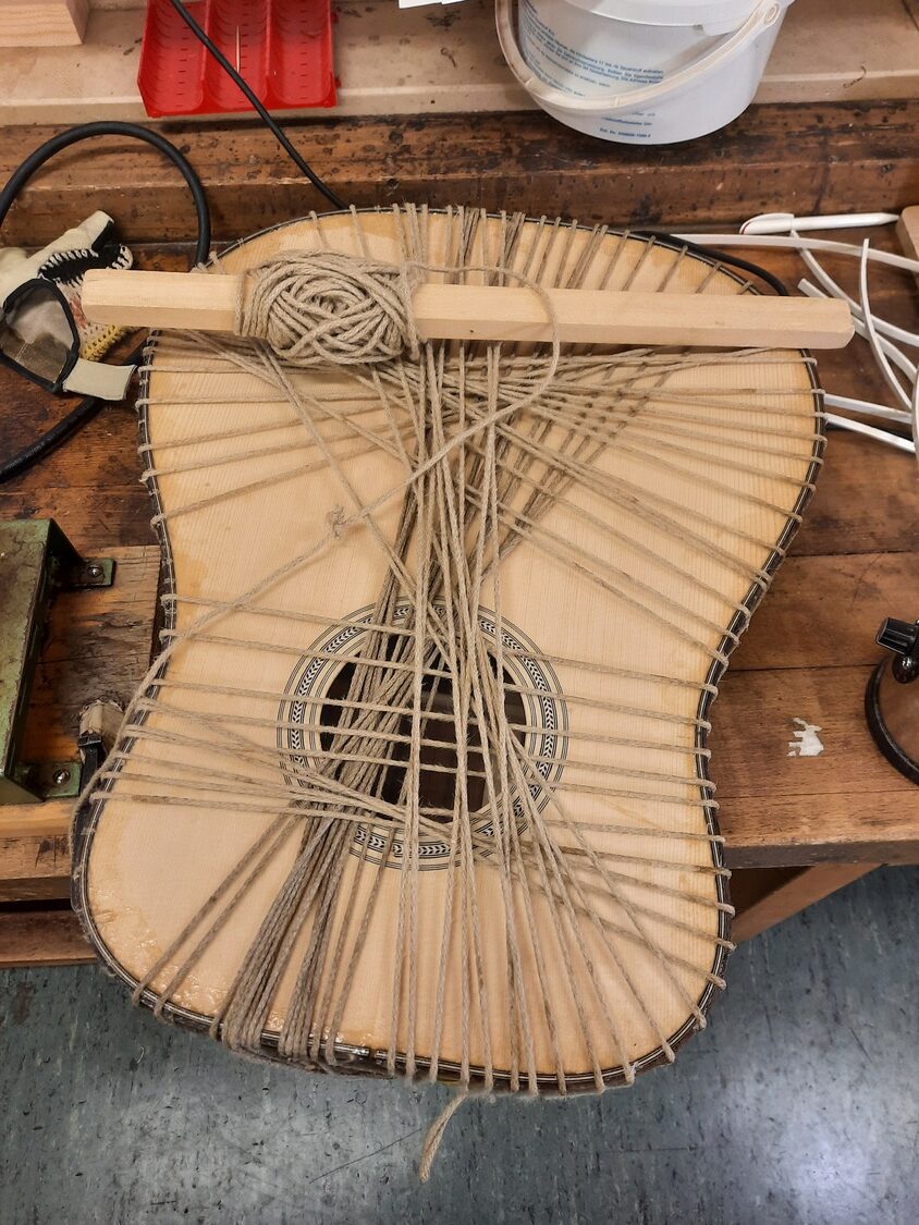 20220210_165152 1 - Deutsche Manufakturenstrasse Gitarrenbody mit Holzstück und Schnur zum Fixieren, auf einer Werkbank mit Werkzeugen und Materialien.