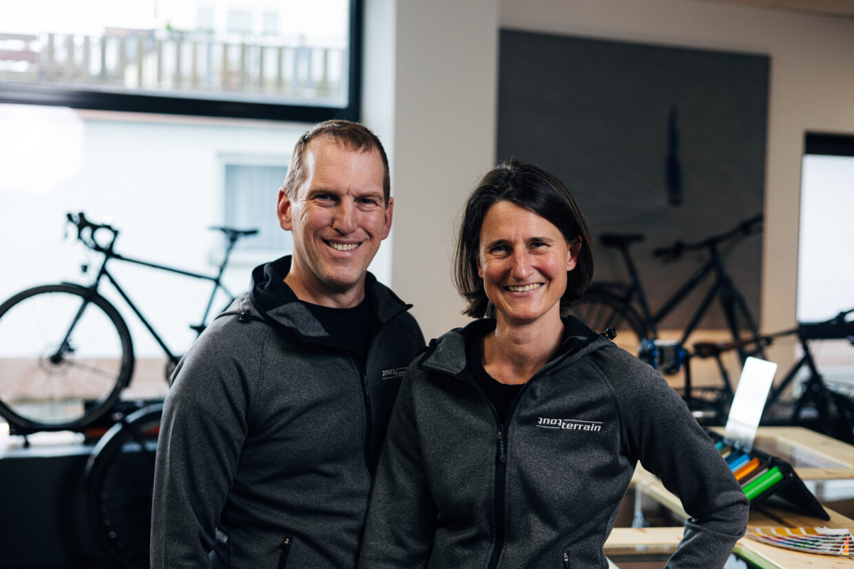 240508_JJANK_ToutTerrain_ManufakturShoot-04811 - Deutsche Manufakturenstrasse Zwei Personen in grauen Jacken lächeln in einem Fahrradgeschäft, im Hintergrund Fahrräder und Arbeitsmaterialien sichtbar.