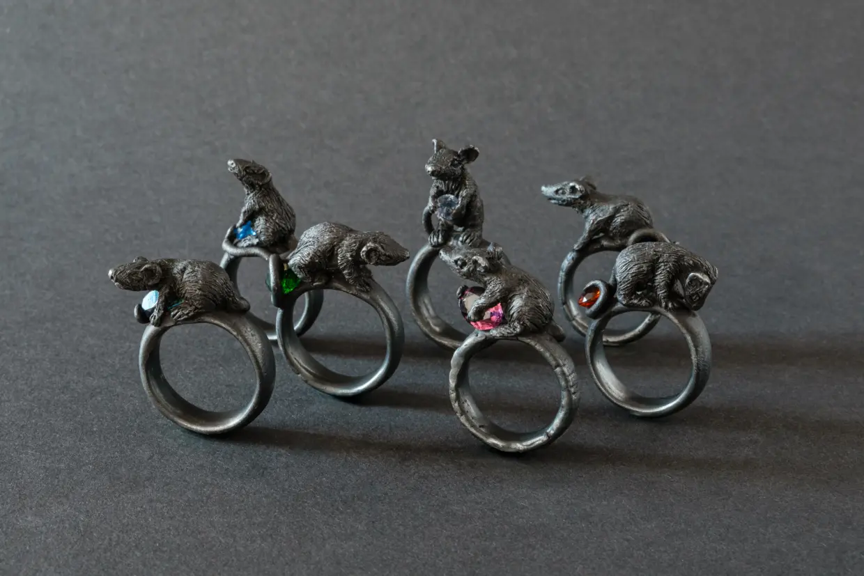 250217_Postkarten-Motive_01_Original - Deutsche Manufakturenstrasse Sechs Ringe mit stilisierten Rattenfiguren, einige mit bunten Edelsteinen, auf einem dunklen Hintergrund.