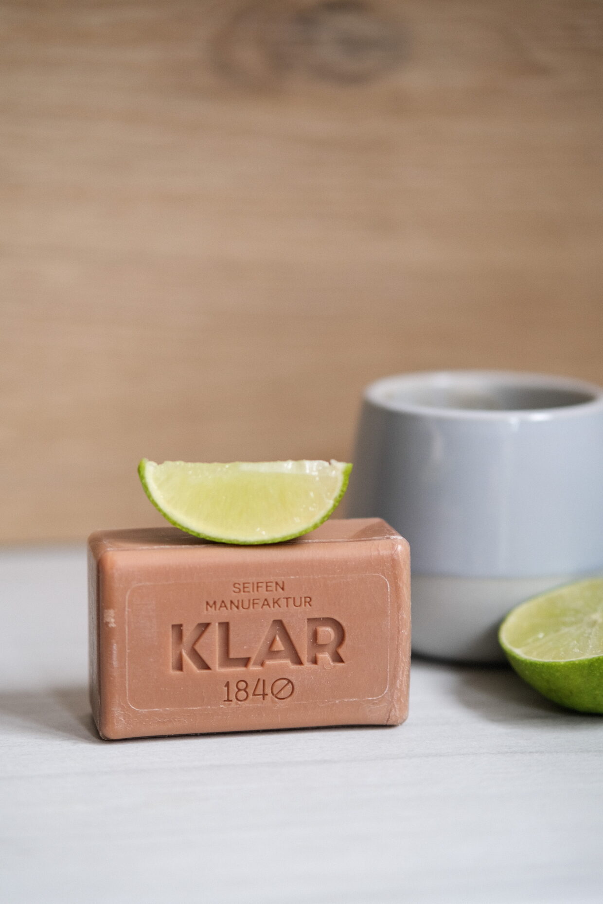 584153692-20240207_up_klar_120 - Deutsche Manufakturenstrasse Buttermilch- und Limettenseife mit einer Limettenscheibe auf einem Holzuntergrund, daneben eine graue Tasse und eine halbe...
