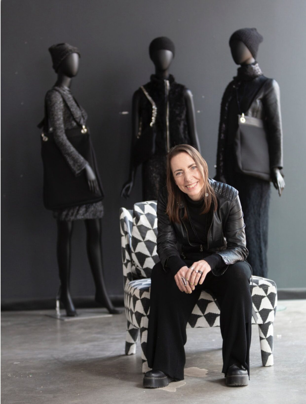 6_Portrait_Monika Assem_1 - Deutsche Manufakturenstrasse Monika Assem sitzt auf einem gemusterten Stuhl vor drei schwarzen Schaufensterpuppen in eleganter Kleidung.
