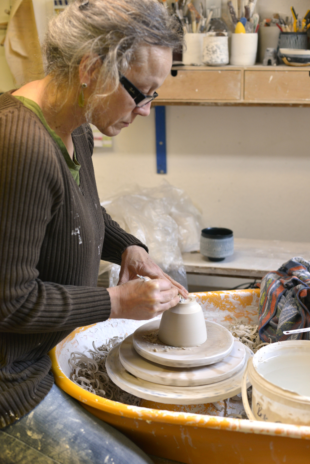 Abdrehen 1 - Deutsche Manufakturenstrasse Frau mit Brille formt einen Tonbecher auf einer Töpferscheibe in einem Atelier, umgeben von Werkzeugen und Materialien.