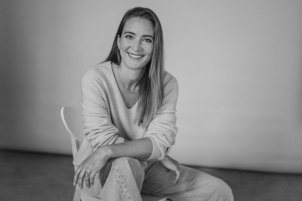 Anna Auras Portrait_sw_Foto Credit Janine Sametzky_10 - Deutsche Manufakturenstrasse Portrait von Anna Auras in Schwarz-Weiß, lächelnd, sitzt entspannt auf einem Stuhl, mit lässiger Kleidung und natürlic...