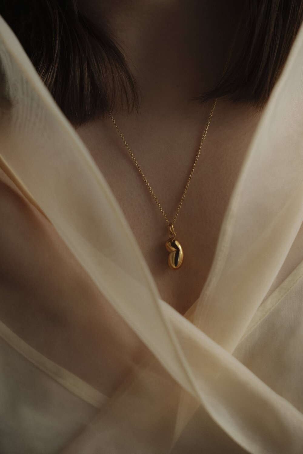 AnnaAuras_Lookbook2021-L_eauKollektion_Foto-JanineSametzky_halskettearp_1 1 - Deutsche Manufakturenstrasse Goldene Halskette mit geschwungenem Anhänger, getragen von einer Person mit schulterlangen Haaren, umgeben von einem tran...