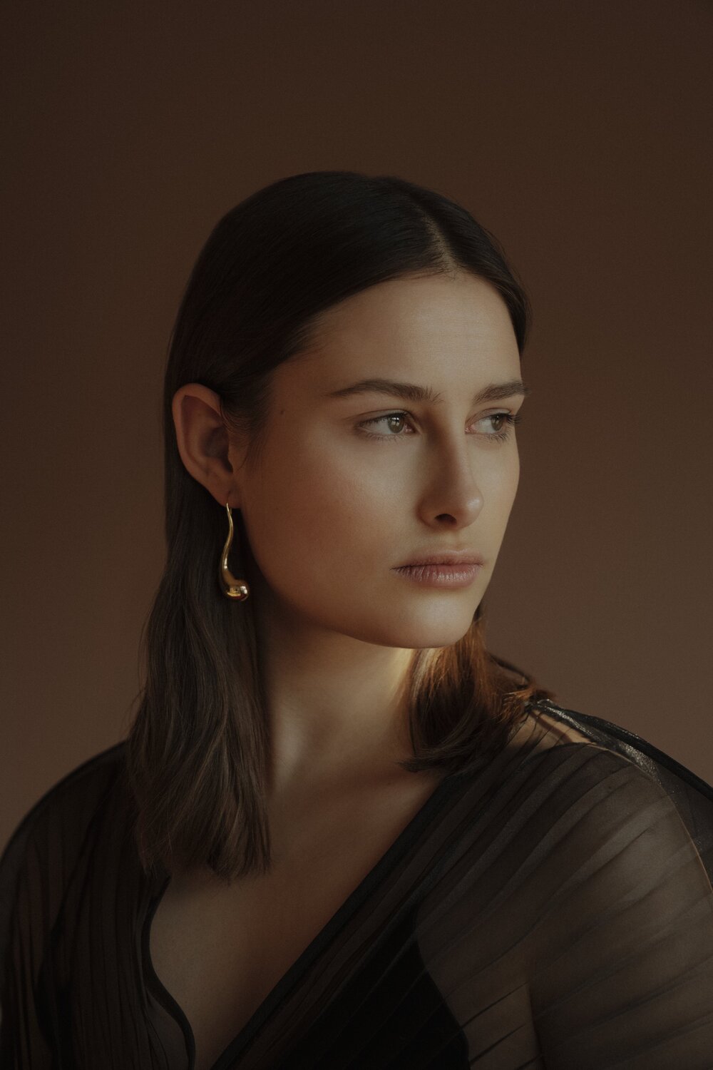 AnnaAuras_Lookbook2021-L_eauKollektion_Foto-JanineSametzky_ohrschmuckdropII_1 2 - Deutsche Manufakturenstrasse Porträt einer Frau mit schulterlangen, glatten Haaren, die goldene Ohrringe trägt, vor einem neutralen Hintergrund.