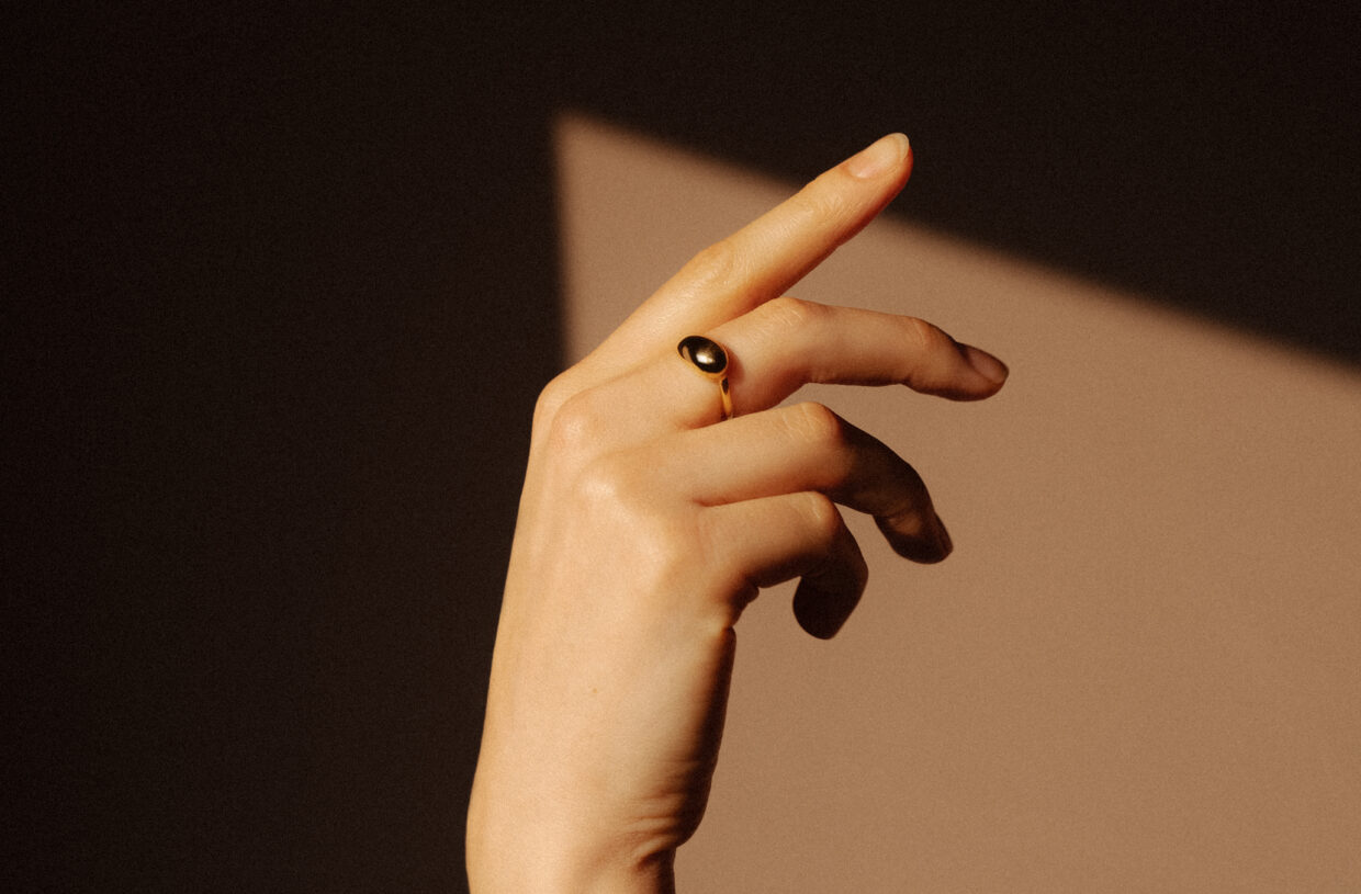 Elegante Hand mit goldenem Ring, vor einem schlichten, schattigen Hintergrund.