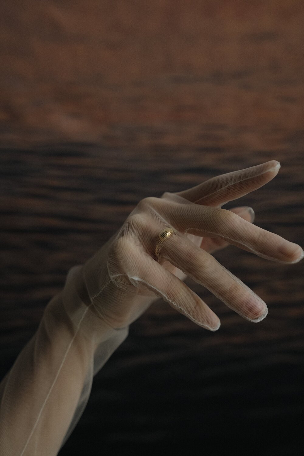 AnnaAuras_Lookbook2021-L_eauKollektion_Foto-JanineSametzky_ringdrop_1 1 - Deutsche Manufakturenstrasse Elegante Hand in transparenter Stoffhandschuh mit goldenem Ring, vor einem verschwommenen, dunklen Hintergrund.