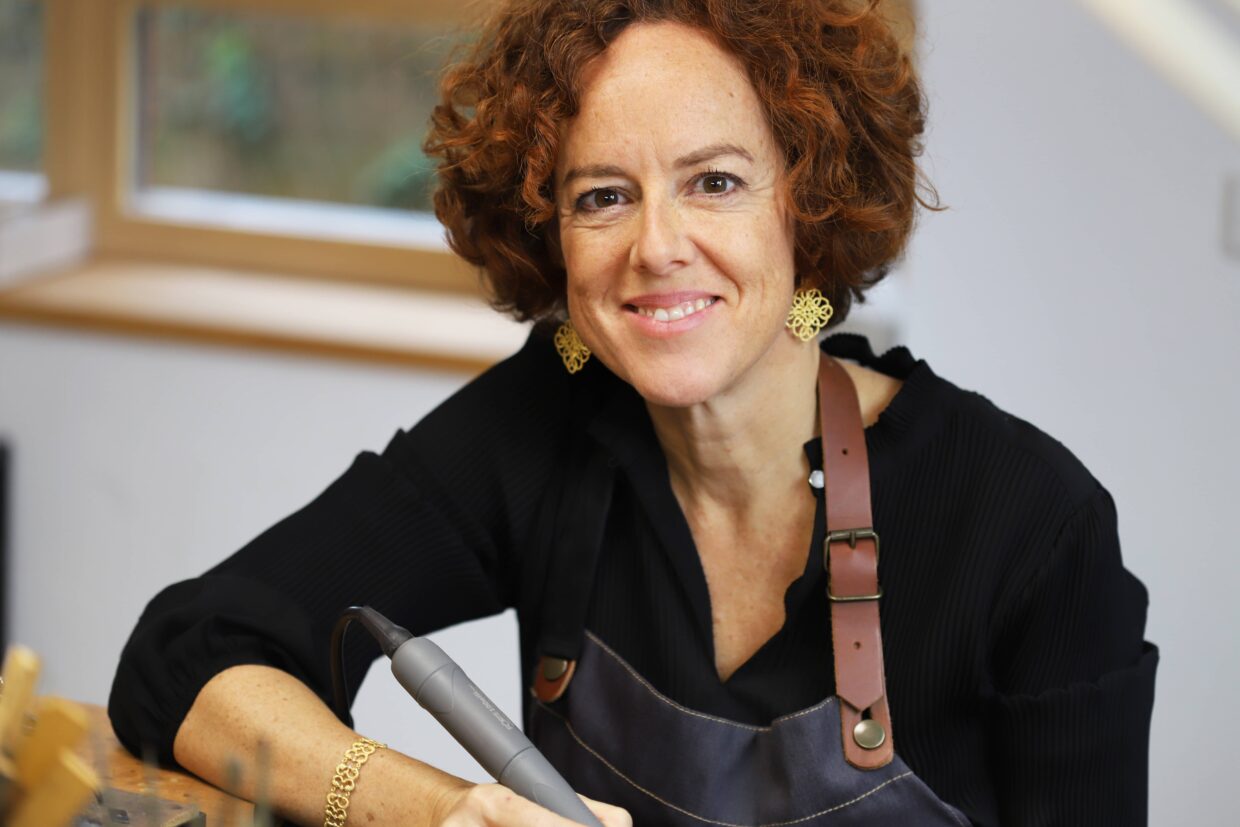 Atelier Adolph-015 - Deutsche Manufakturenstrasse Frau mit lockigem, rotem Haar und Ohrhängern, sitzt in einem Atelier und lächelt, während sie ein Werkzeug in der Hand ...