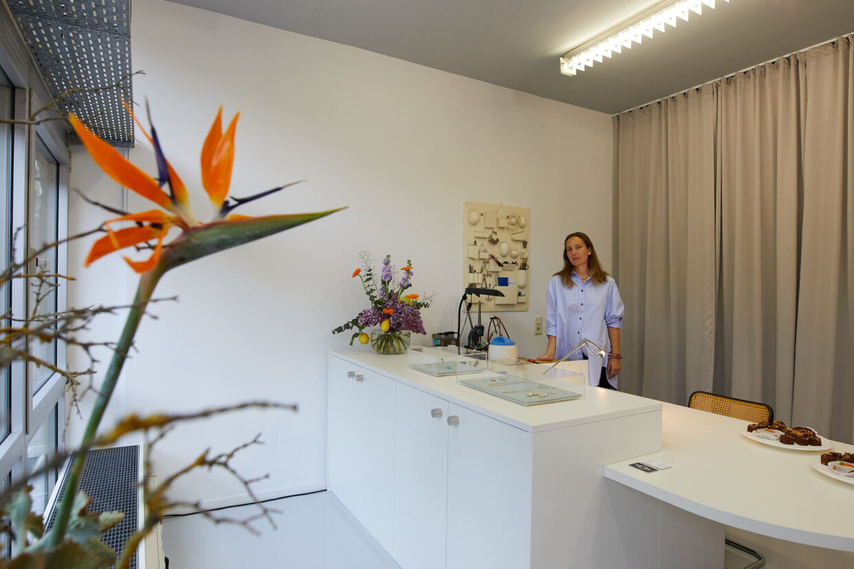 Atelier_Annaauras_2022 1 - Deutsche Manufakturenstrasse Helle Atelier-Umgebung mit einer Frau in blauer Bluse, Blumenarrangement und einem Tisch mit Snacks.