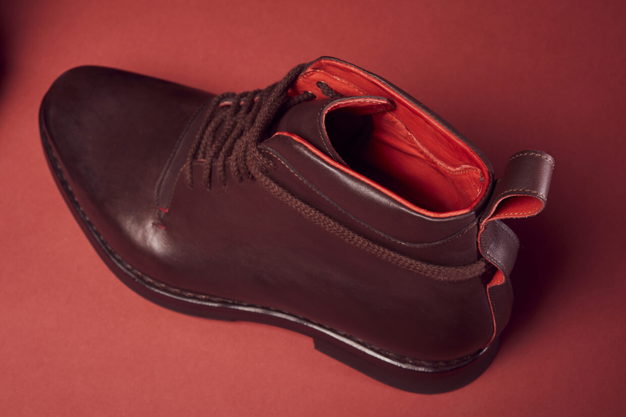 BB2511 1 - Deutsche Manufakturenstrasse Braune Lederstiefelette mit roten Innenfutter und Schnürsenkeln, auf rotem Hintergrund platziert.