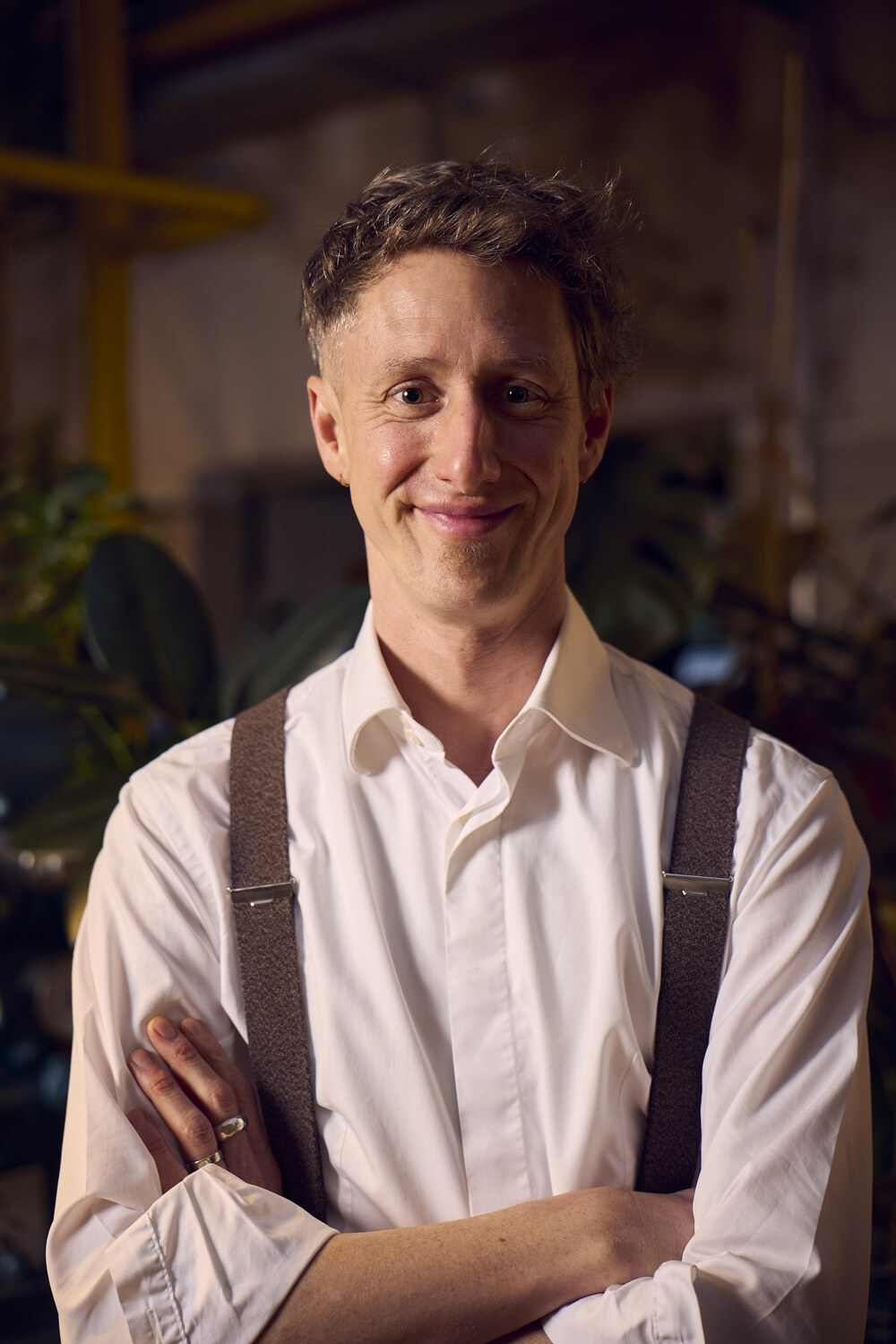 BB_Family2025_128 1 - Deutsche Manufakturenstrasse Lächelnder Mann mit kurzen Haaren, weißem Hemd und Hosenträgern, steht vor einer grünen Pflanzenkulisse.