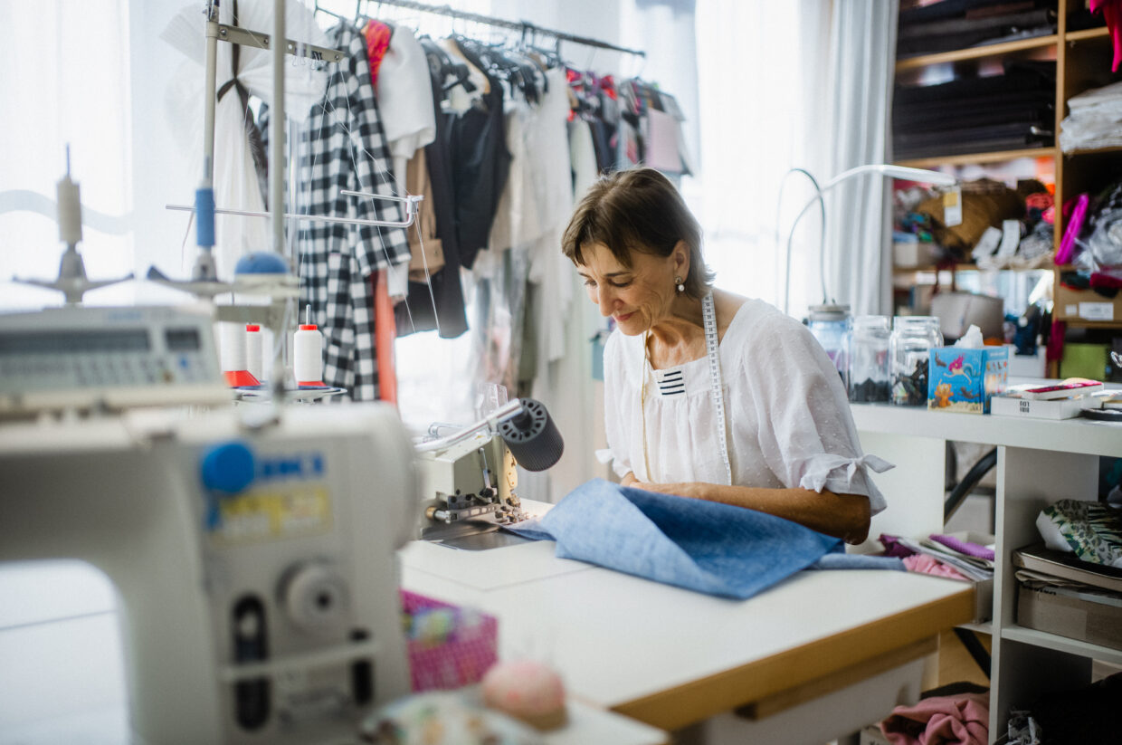 Bild 3 1 - Deutsche Manufakturenstrasse Frau näht an einer Nähmaschine in einem hellen Atelier, umgeben von Stoffen und Kleidung.