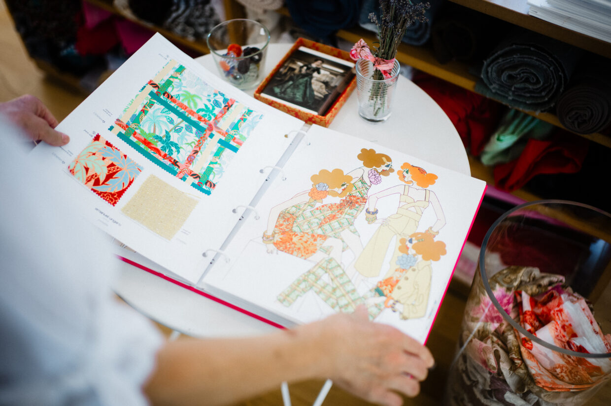 Bild 4 1 - Deutsche Manufakturenstrasse Person blättert durch ein Designbuch mit bunten Illustrationen und Stoffmustern auf einem Tisch.