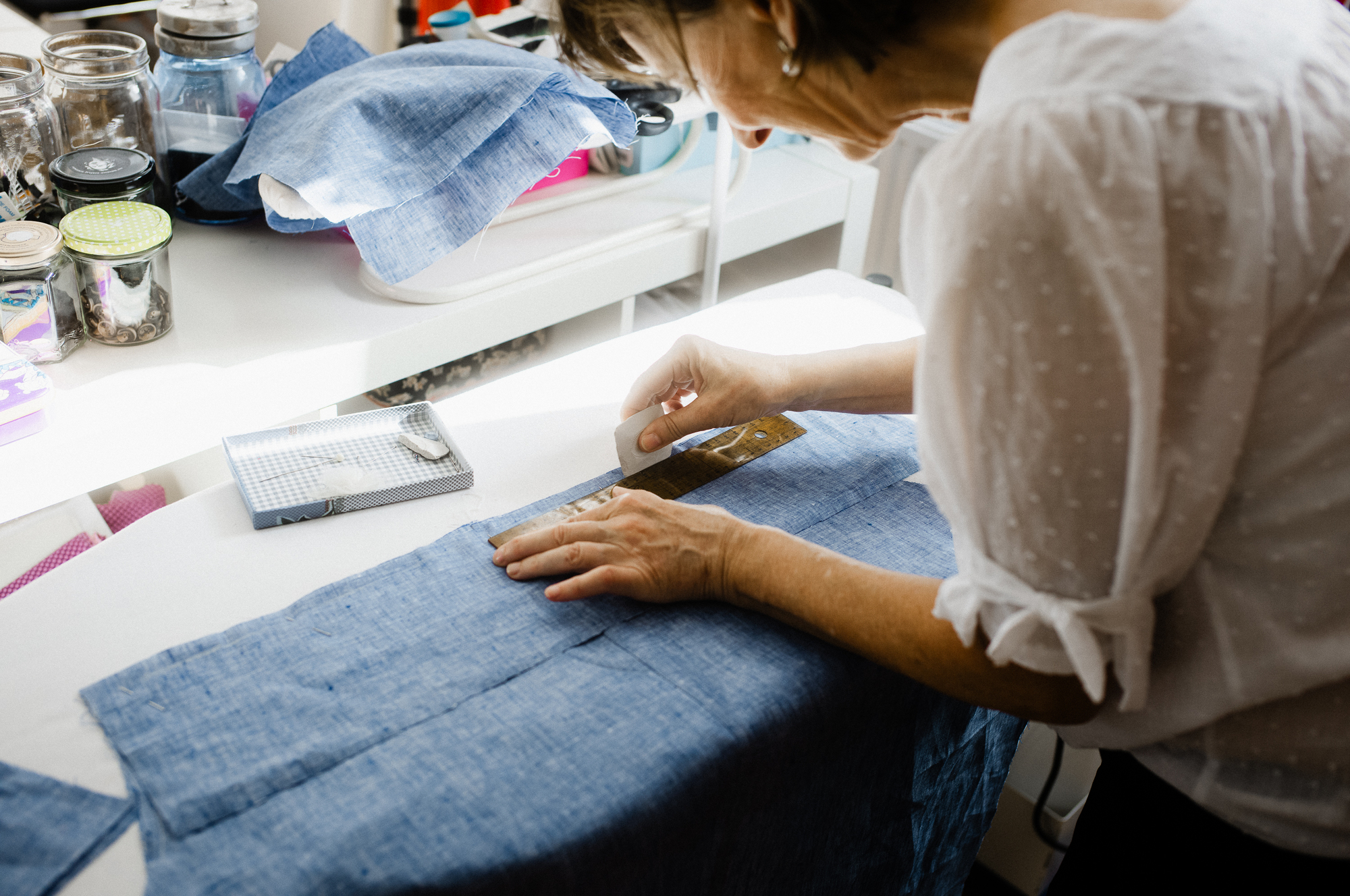 Bild 5 1 - Deutsche Manufakturenstrasse Frau misst blauen Stoff mit einem Lineal auf einem Tisch, umgeben von Nähutensilien und Gläsern.