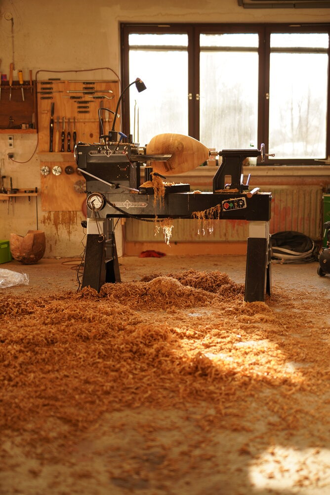 DSC04069 1 - Deutsche Manufakturenstrasse Holzwerkstatt mit einer Drehmaschine, umgeben von Holzspänen und Tageslicht, das durch ein Fenster strömt.