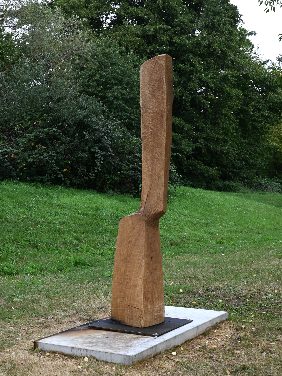 Douma_Kleine Drehung 1 - Deutsche Manufakturenstrasse Holzskulptur mit einer schlanken, vertikalen Form, auf einem grauen Sockel, umgeben von Grünflächen.