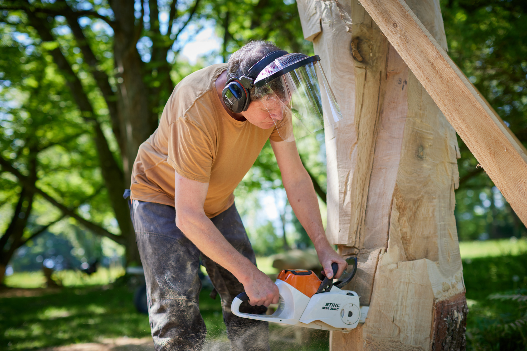 Douma_Porträt - Deutsche Manufakturenstrasse Mann mit Gehörschutz und Gesichtsschutz schneidet mit einer Kettensäge einen Baumstamm im Freien.