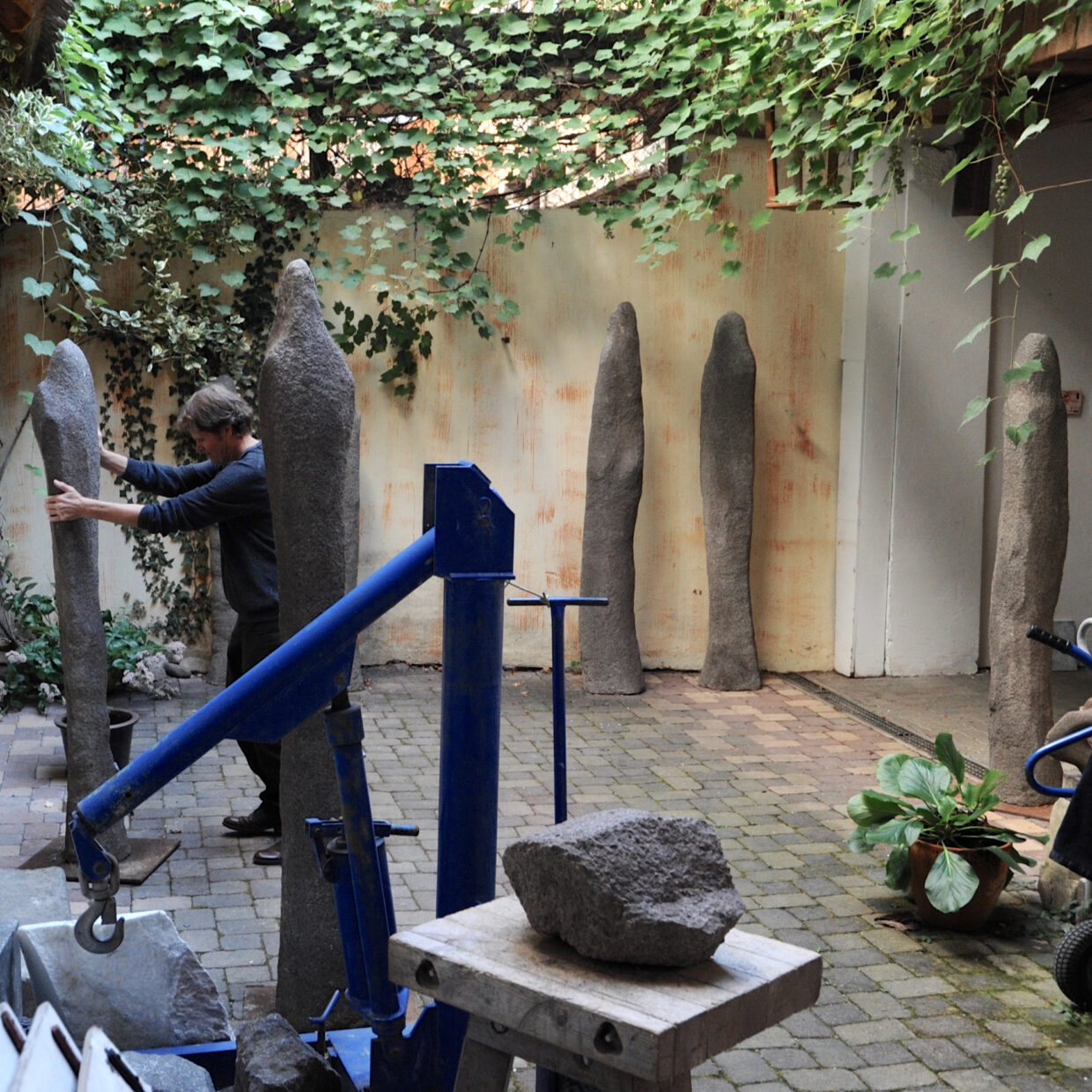 Douma_Werkstatt 1 - Deutsche Manufakturenstrasse Ein Künstler in einer Werkstatt arrangiert mehrere steinerne Skulpturen in einem begrünten Innenhof.