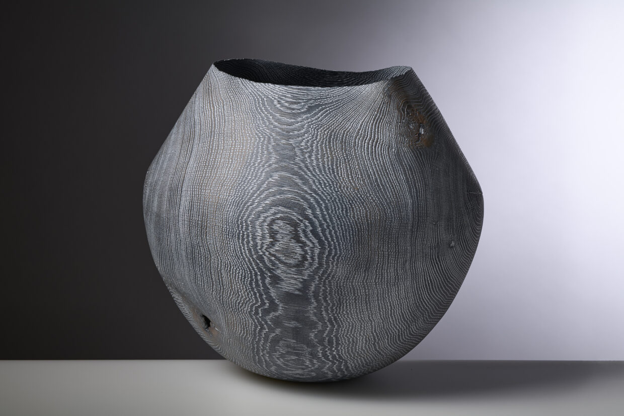 GLO 22 1 - Deutsche Manufakturenstrasse Graue, ovale Holzskulptur mit ausgeprägter Maserung und unregelmäßiger Öffnung, auf einem hellen Untergrund.