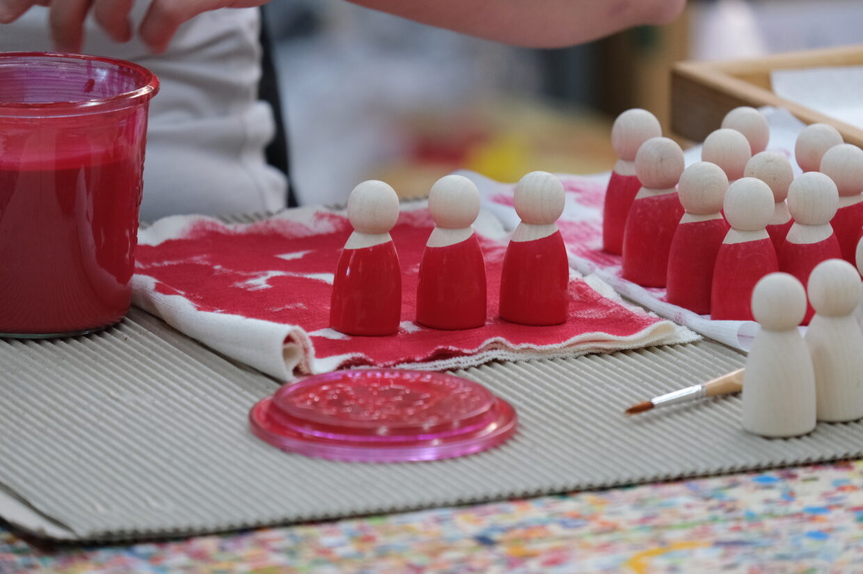 GRIMMS_Regenbogen_Freunde_Handbemalen 1 - Deutsche Manufakturenstrasse Handbemalte Holzfiguren in Rot stehen auf einem Tisch, daneben ein Glas mit roter Farbe und ein Pinsel.