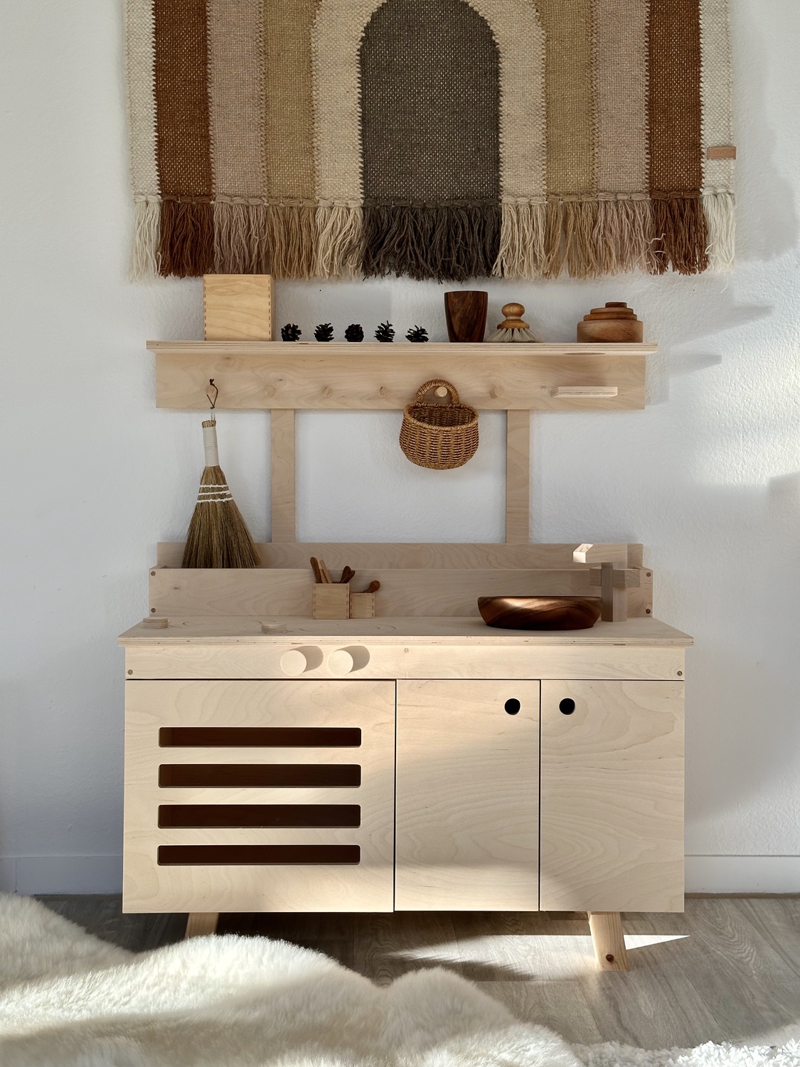 GRIMMS_UMI_SPIELKUECHE 1 - Deutsche Manufakturenstrasse Holzküche mit offenen Regalen, dekoriert mit Körben und Holzspielzeug, vor einem gewebten Wandteppich.