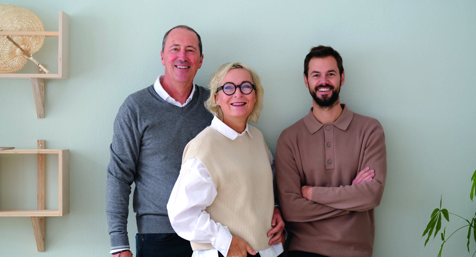 GRIMMS_vl_SvenGrimm_ElkeGrimm_MarcoStuber 1 - Deutsche Manufakturenstrasse Drei Personen stehen lächelnd vor einer hellen Wand: ein Mann links, eine Frau in der Mitte und ein Mann rechts.