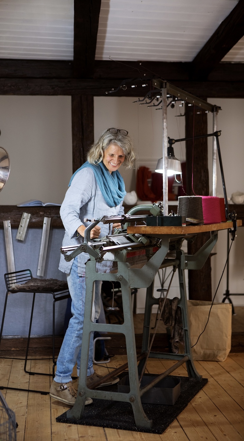 Grieb Uschi Strickmaschine Foto Joss Andres 1 - Deutsche Manufakturenstrasse Uschi Grieb steht an einer Strickmaschine in einem Atelier, umgeben von Garn und Werkzeugen.