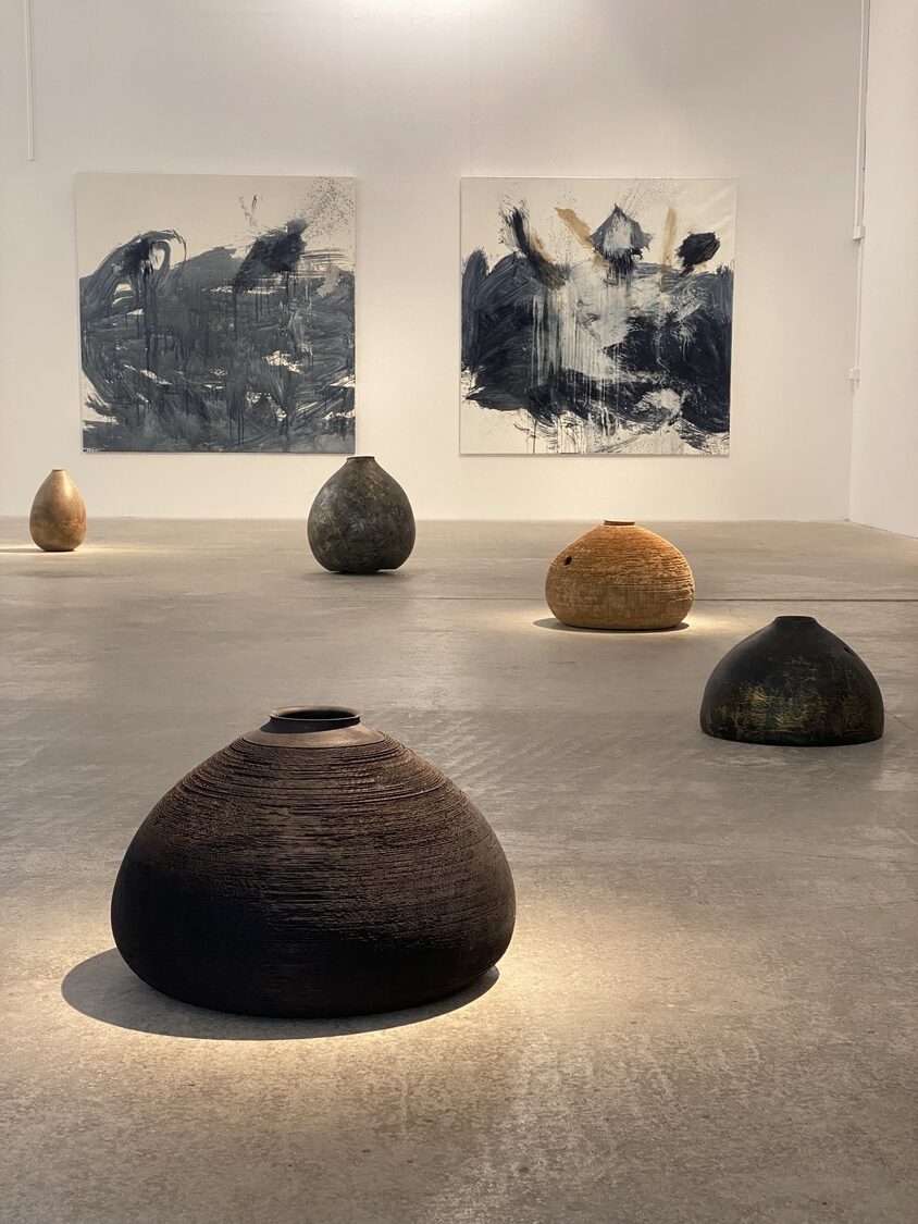 IMG_0981 1 - Deutsche Manufakturenstrasse Dunkle, runde Keramiken im Vordergrund, umgeben von hellen, abstrakten Wandbildern in einer Galerie.