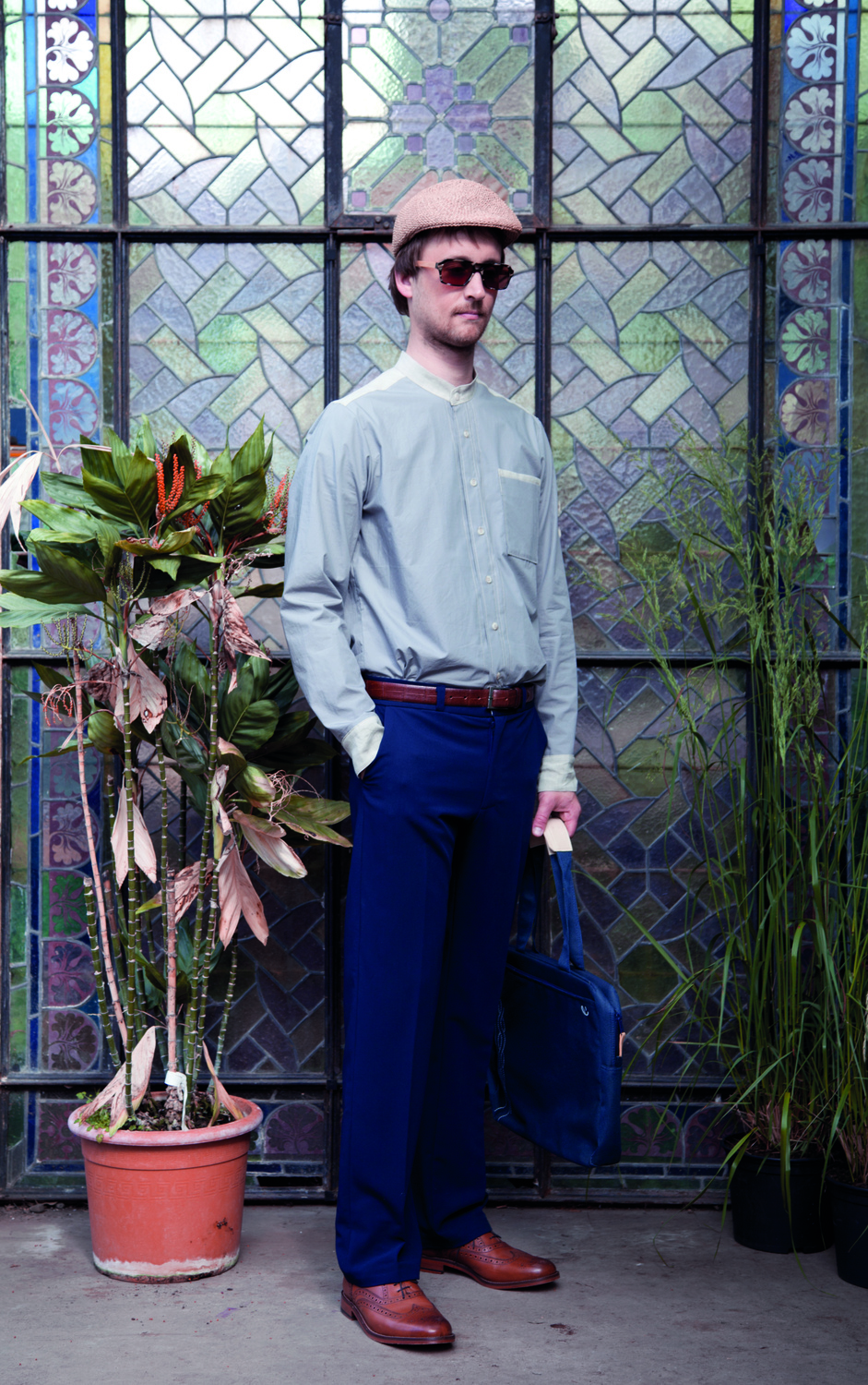 IMG_6794 1 - Deutsche Manufakturenstrasse Mann in modischer Kleidung mit Sonnenbrille und Hut steht vor buntem Glasfenster, umgeben von Pflanzen.