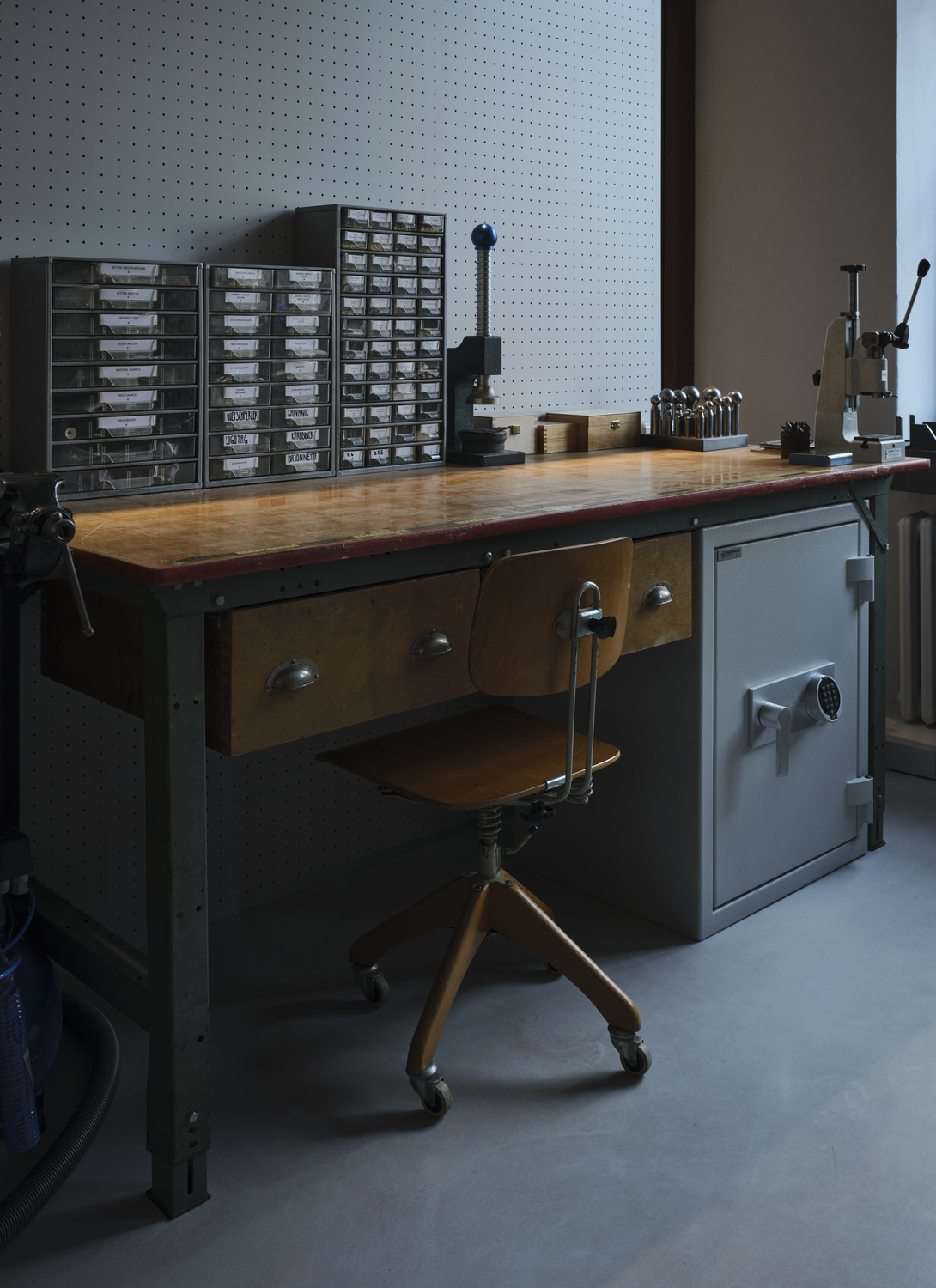 JJ_Showroom-Manufaktur_Feb2023_3535++ 1 - Deutsche Manufakturenstrasse Werkbank mit Holzplatte, Stuhl und Werkzeugaufbewahrung in einem hellen Raum. Metallregal mit Schubladen im Hintergrund.