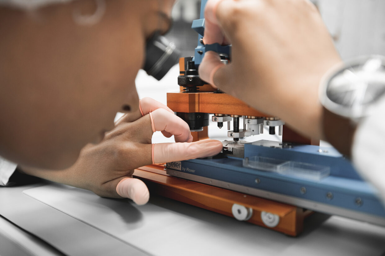 Junghans production 3 1 - Deutsche Manufakturenstrasse Detailaufnahme einer Person, die präzise an einer Uhrmachermaschine arbeitet, mit Fokus auf die Hände und das Werkzeug.