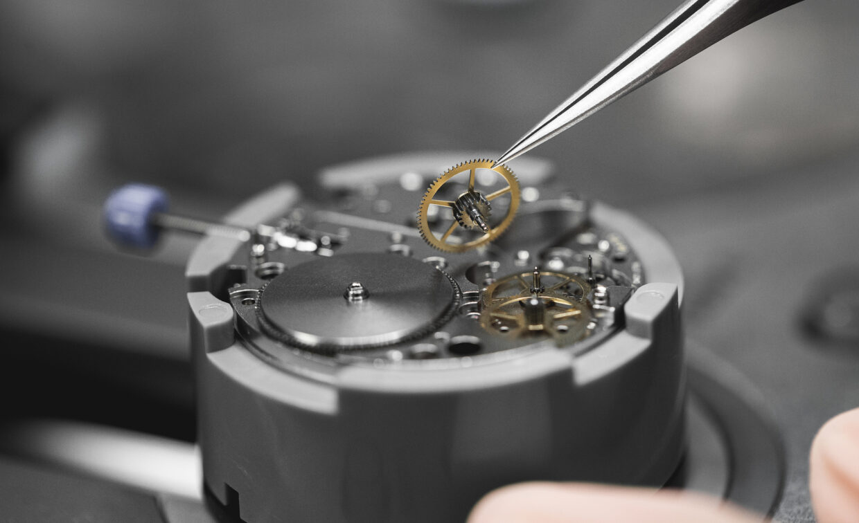 Junghans_assembly mechanical calibre 1 - Deutsche Manufakturenstrasse Detailansicht einer Uhrwerk-Assemblierung mit einer Pinzette, die ein Zahnrad hält, auf einem grauen Untergrund.