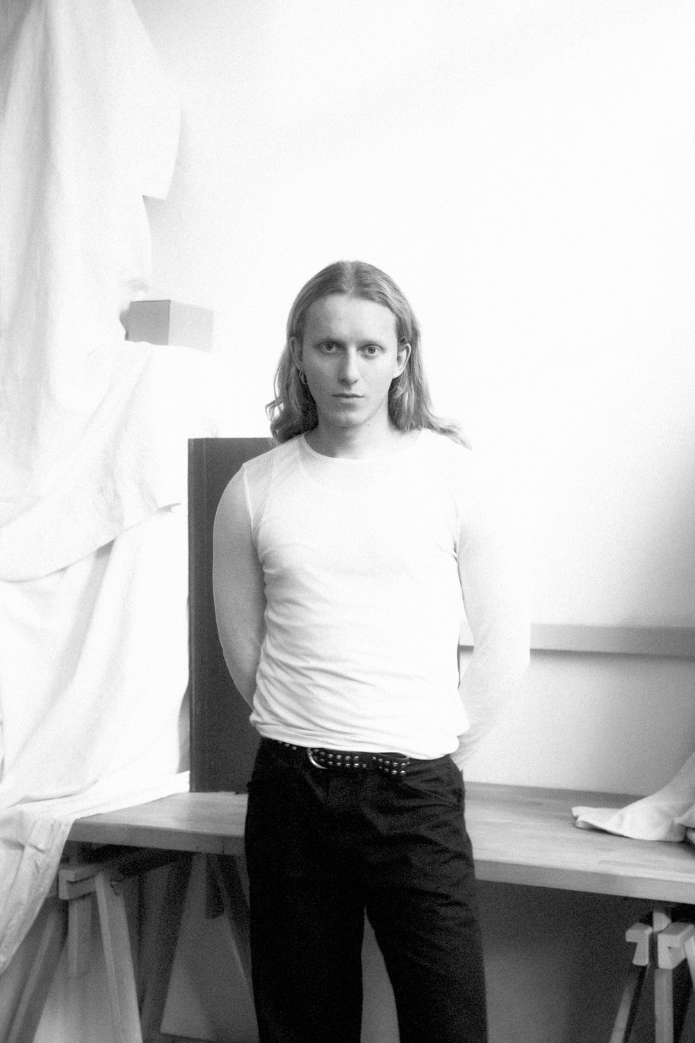 Portrait 1 - Deutsche Manufakturenstrasse Junger Mann mit langen Haaren in einem weißen Shirt, steht in einem Atelier mit hellem Hintergrund.