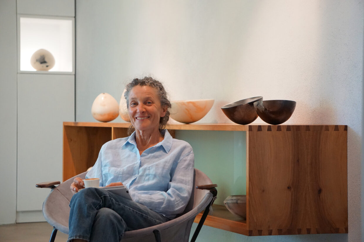 Portrait im Ausstellungsraum 1 - Deutsche Manufakturenstrasse Lächelnde Frau sitzt in einem Ausstellungsraum, hält eine Tasse, hinter ihr sind keramische Objekte auf einem Regal.